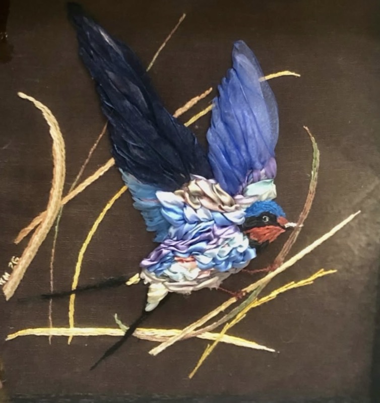 A swallow with its blue wings raised, worked in silk ribbon among fine reeds on a dark ground